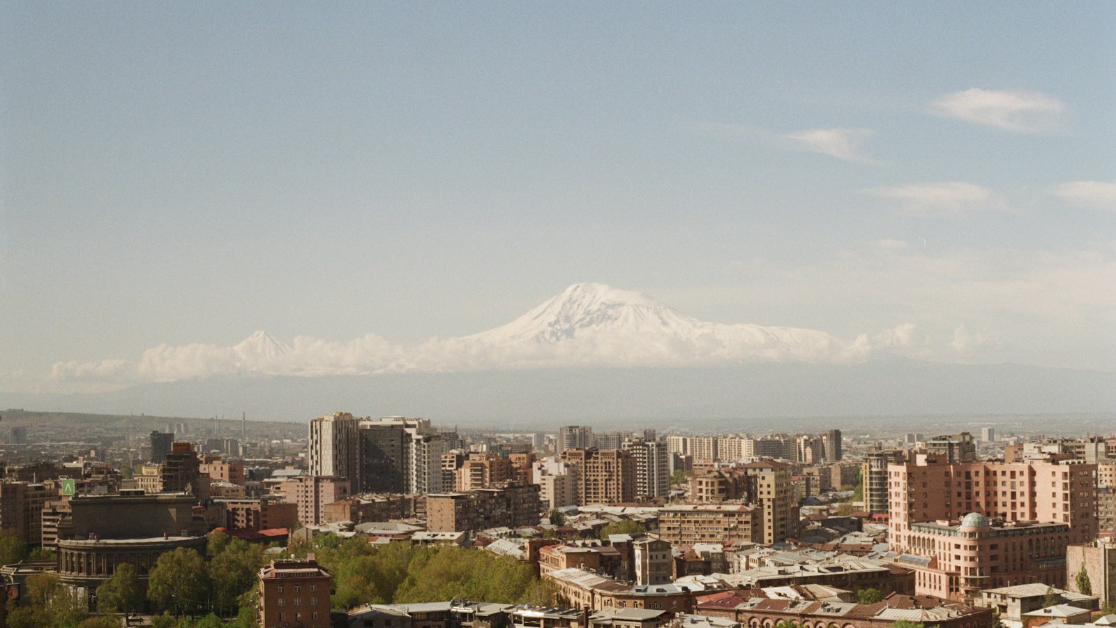 Երևանի համայնապատկերը կապույտ երկնքի տակ, իսկ քաղաքի հետևում բարձրանում է Արարատի ձյունածածկ գագաթը։