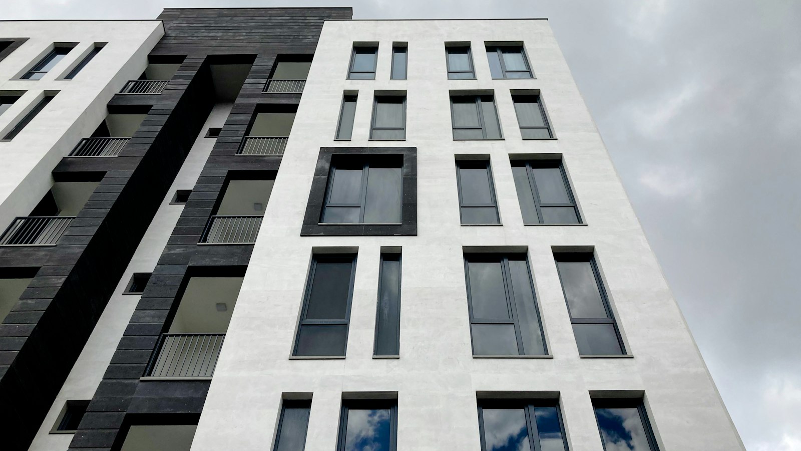 Looking up at a modern white and dark-grey apartment block with balconies in Yerevan under a cloudy sky.